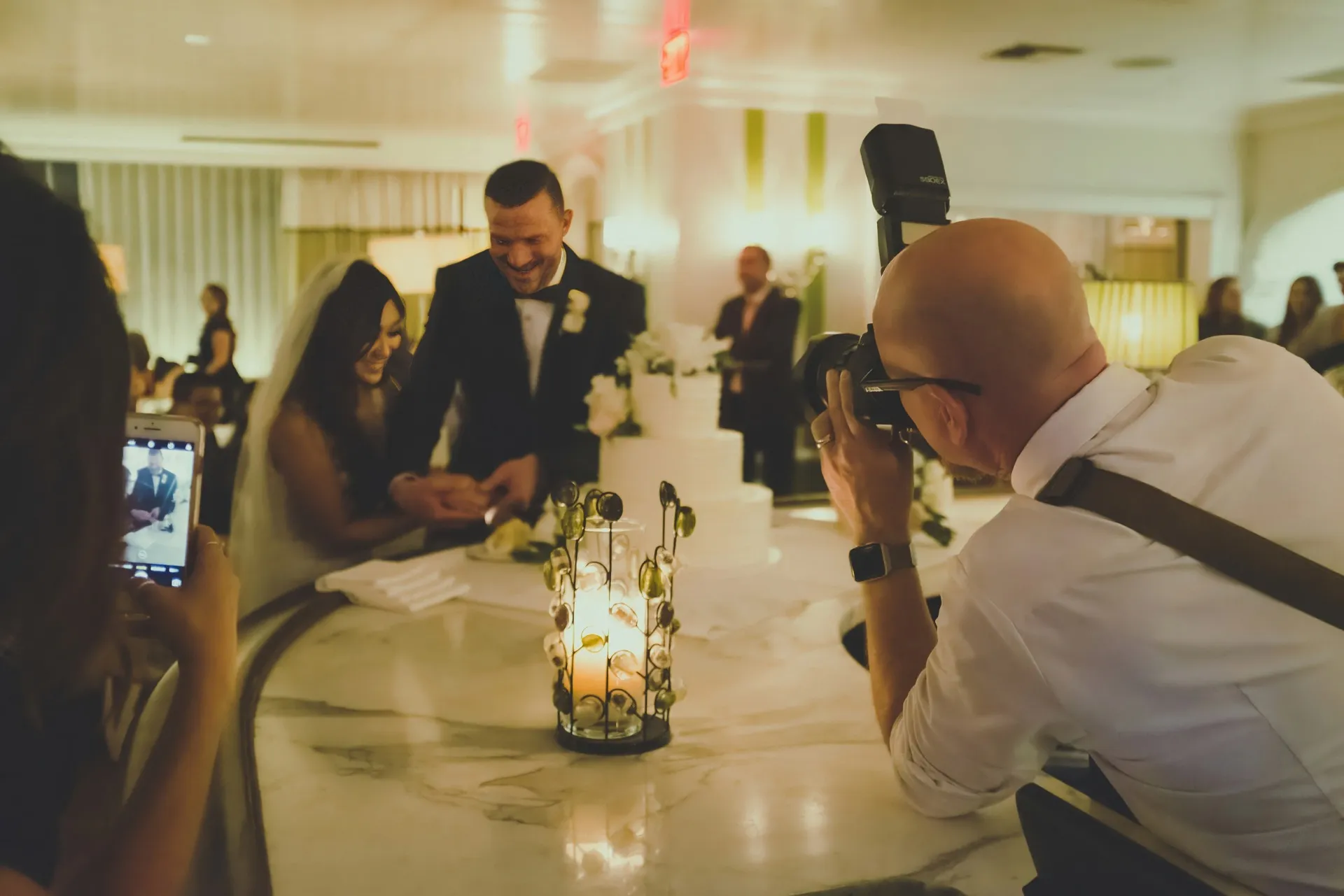 A wedding videographer leaning into frame while recording a bride and groom cutting their wedding cake.